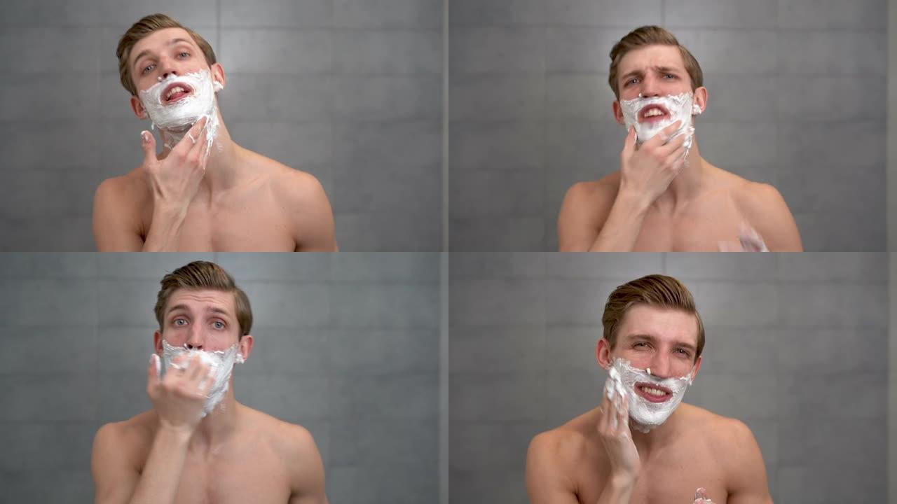 英俊的未刮胡子的年轻人在脸上涂抹保湿剃须泡沫，看着相机。快乐男人在浴室在家进行日常个人护理的肖像