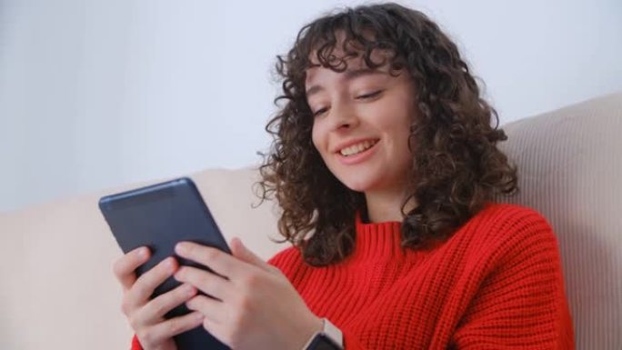 快乐的白人女人使用平板电脑，露齿微笑。快乐的卷发年轻女性在现代便携式设备上浏览快速5g互联网。可爱的