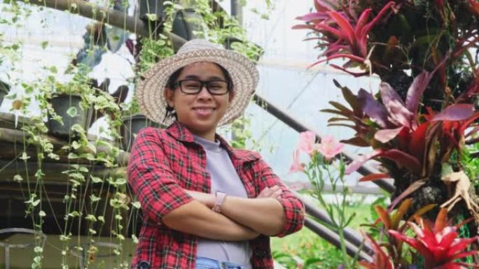 一个微笑的园丁女人的肖像，双臂交叉在温室中的花卉植物上站着。在花园工作的年轻女企业家。小企业主的启动