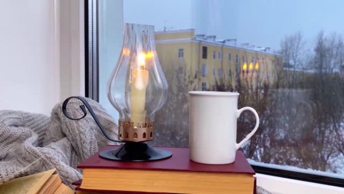 咖啡杯，烟熏，蜡烛燃烧和窗台上的书，冬天的早晨或傍晚，外面下雪，风移动页面。选择性聚焦