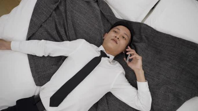 顶视图不堪重负的亚洲商人躺在床上拿着电话在家里的卧室里精力不足地聆听和回复