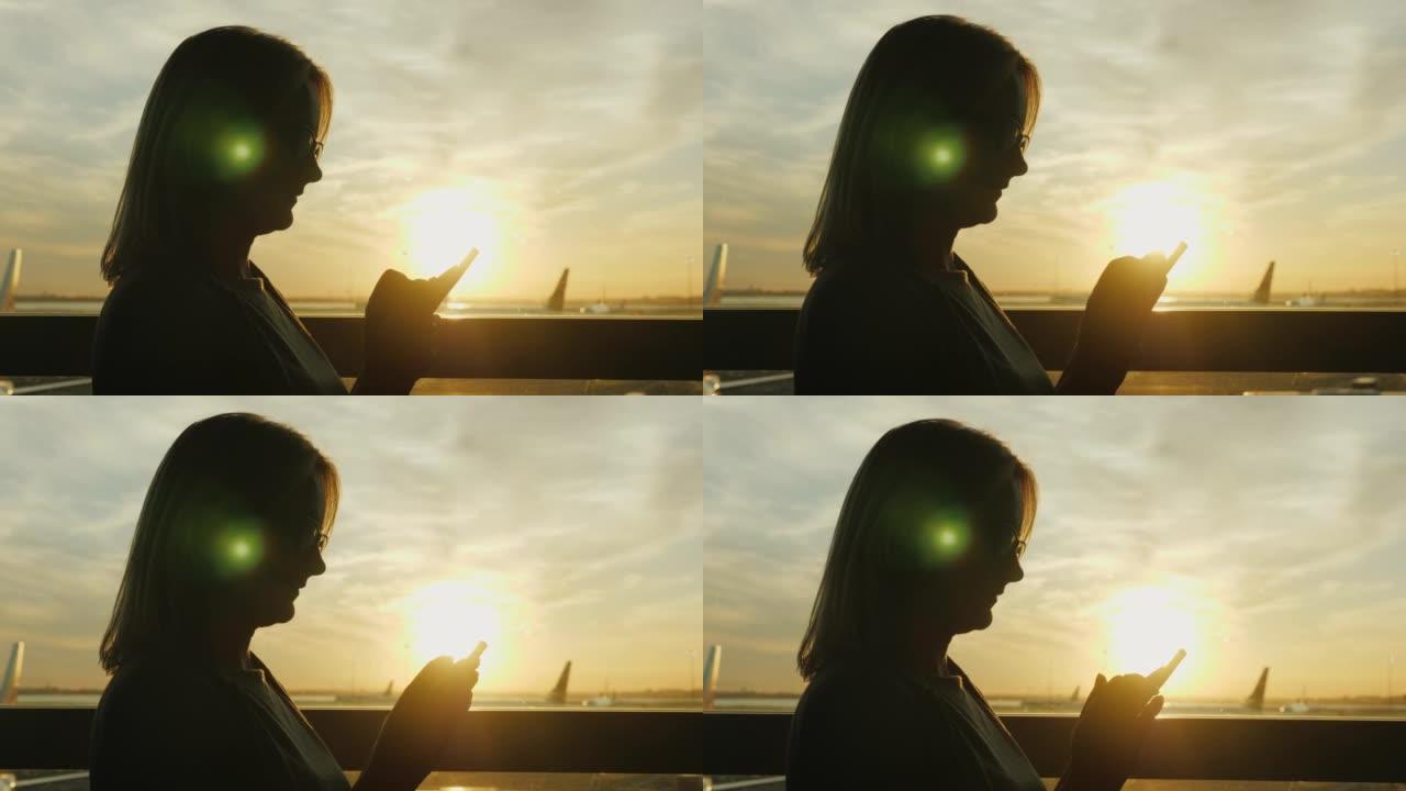 机场航站楼一个大窗户上的一名妇女的剪影。使用智能手机
