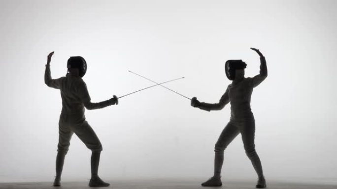 两名女剑客决斗的剪影。运动员越过剑杆并进行攻击。女孩们在聚光灯下的白色背景下的黑暗工作室里摆姿势。慢