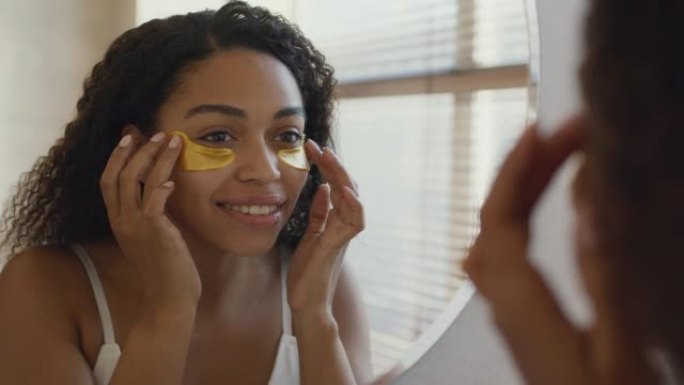 年轻漂亮的非洲裔美国妇女在脸上涂抹抗水肿胶原蛋白贴片，在浴室照镜子