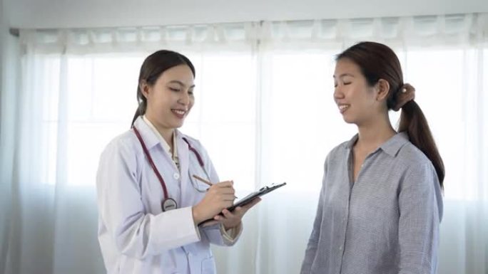 4K 50fps，白人亚裔女医生站在医院检查室对一名身穿长袖衬衫的长发亚裔患者做笔记 ....