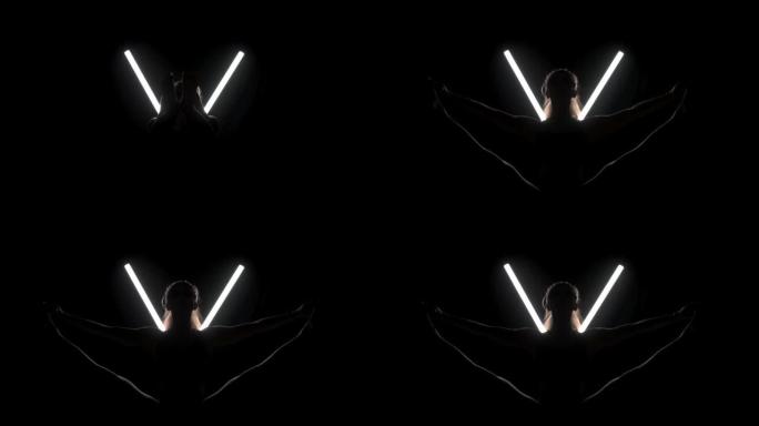 练习瑜伽姿势的健康女性。剪影女孩在黑色背景下的工作室里做运动，带v形白色发光二极管灯管。无压力内部平