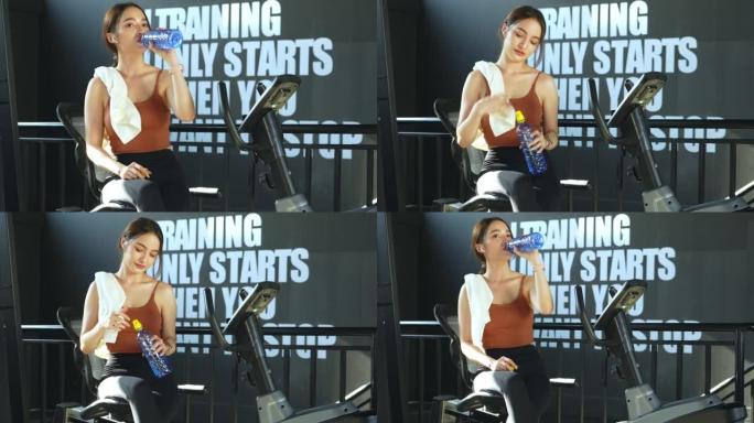 一名亚洲妇女在健身房休息，在健身在线小时用减肥锻炼机锻炼后，用毛巾擦干脸，喝矿泉水