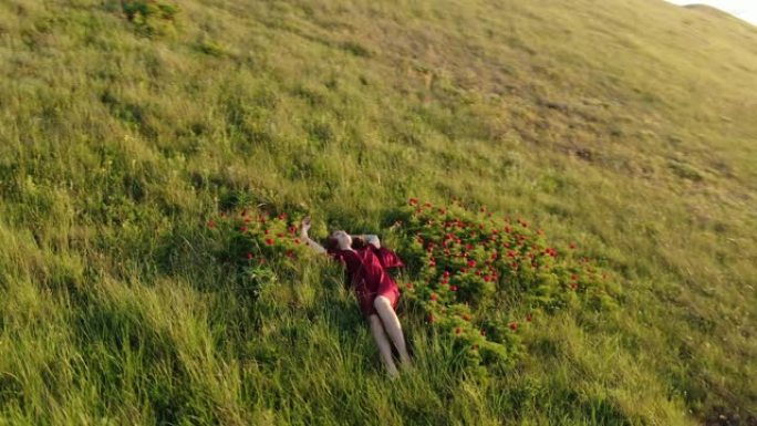 慢动作。鸟瞰图，一个穿着红色连衣裙的年轻美女坐在红色野生牡丹的草地上