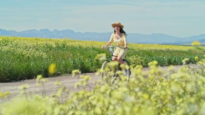 4k视频片段，一名年轻女子在乡村花田旁的一条土路上骑自行车