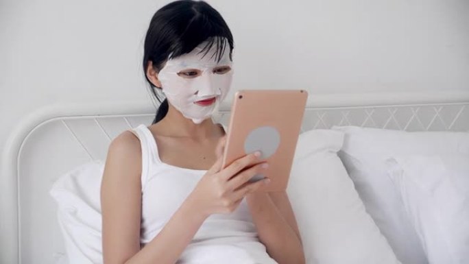 美丽的年轻亚洲妇女与床单面膜坐在卧室的床上看平板电脑，美容女孩应用面膜在家里的皱纹护肤，皮肤护理，健