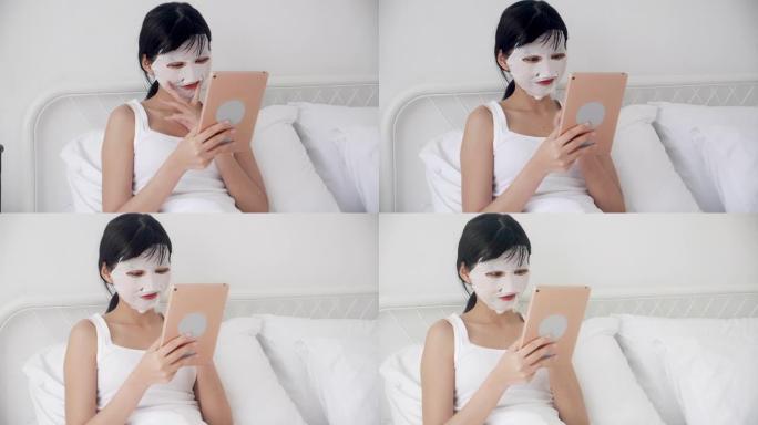 美丽的年轻亚洲妇女与床单面膜坐在卧室的床上看平板电脑，美容女孩应用面膜在家里的皱纹护肤，皮肤护理，健