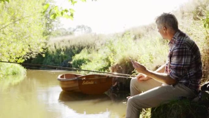 成熟的男人在阳光明媚的夏日河钓鱼和使用智能手机