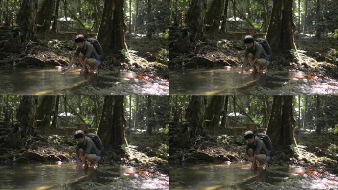 背着背包的女徒步旅行者在热带森林树荫下的小溪中洗手。