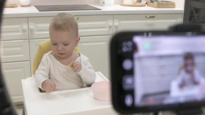 父母在家里高脚椅上的孩子的智能手机上录制视频，用手机和铃声录制一岁大的男婴水果的照片视频。