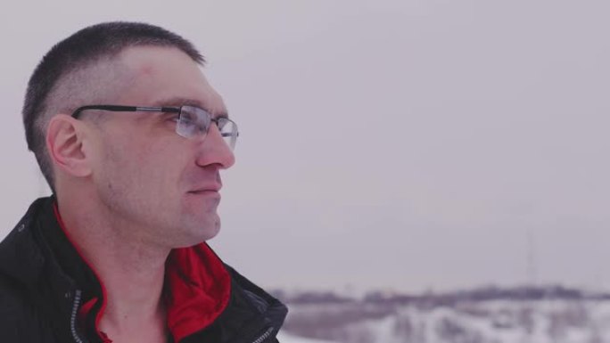 一个戴眼镜的成年男子若有所思地望着远方，冬天在下雪的天气里站在大街上。穿着外套和眼镜的成年男性