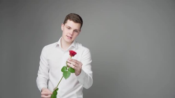 一个穿着白色衬衫的年轻人，在灰色的背景上，献上一朵红玫瑰。