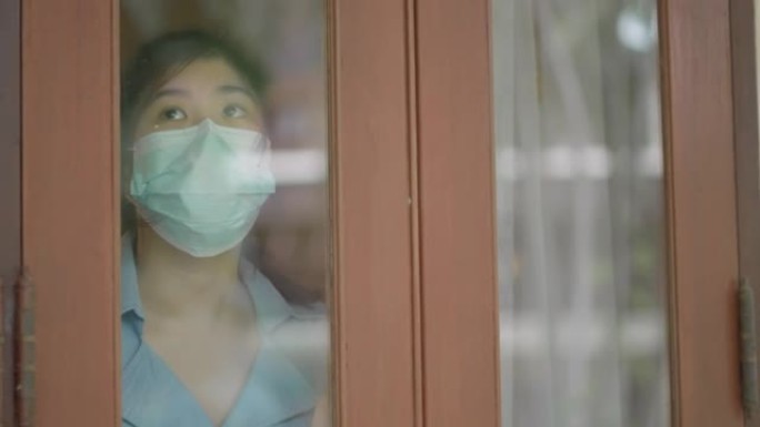 戴口罩的亚洲女孩在屋内用悲伤的眼神看着外面
