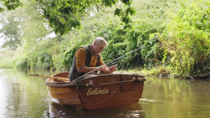 成熟的人从河上的木船钓鱼和使用智能手机