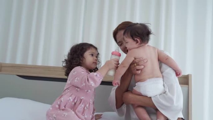 亚洲单身母亲在卧室照顾孩子，小女孩帮助奶瓶喂养婴儿