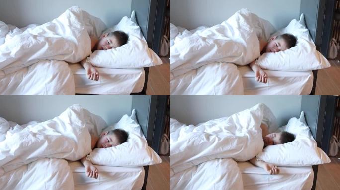 昏昏欲睡的蹒跚学步的男孩躺在床上，穿着白色床上的衣服哭泣。卧室里累了的孩子。