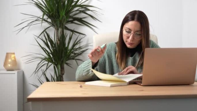 年轻的企业家女性，戴眼镜在笔记本电脑上工作，在舒适的办公室里在笔记本上写笔记