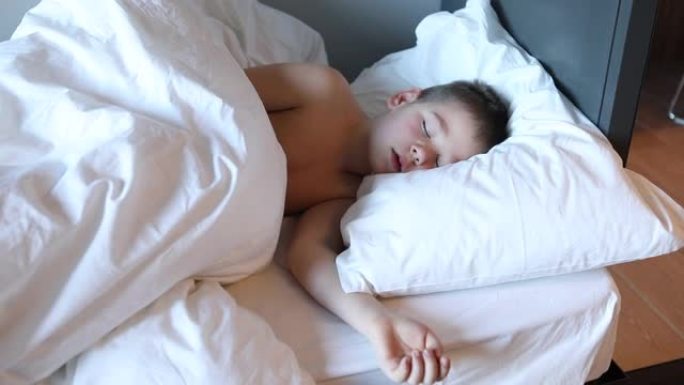 悲伤的蹒跚学步的男孩躺在床上，穿着白色的床上用品哭泣。卧室里累了的孩子。7岁的孩子躺在床上，昏昏欲睡