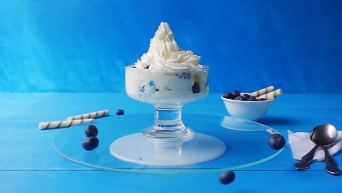 蓝莓落入装有冰淇淋的玻璃杯中，在蓝色背景下的木制桌子上的托盘上旋转。具有速度斜坡效果的4k慢动作视频