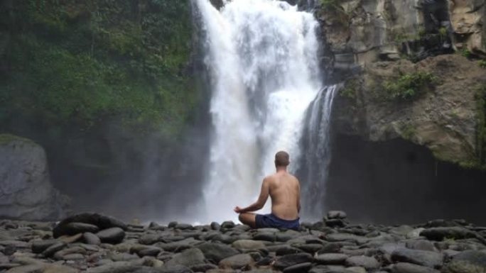 冷静集中的人沉思，正念在雨林丛林中巨大的坚固瀑布前。宁静使男人充满了自然的力量。一个人充满了水的能量