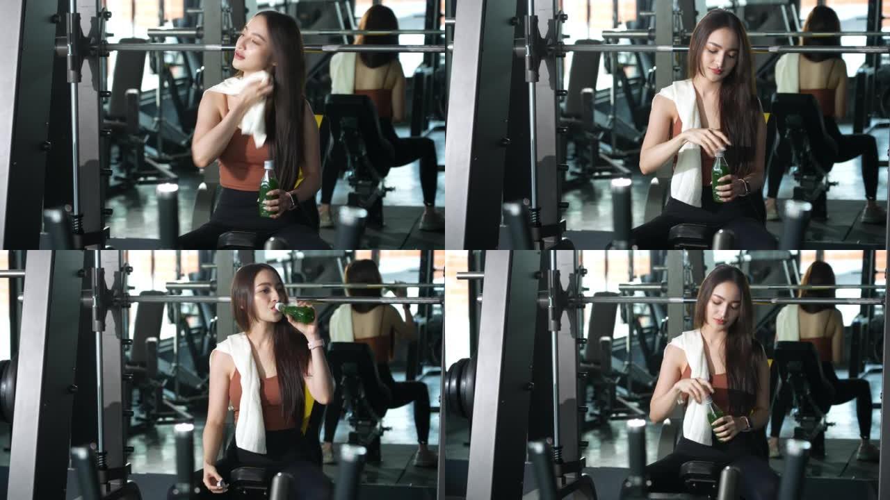 一名亚洲妇女在健身房休息，在健身时间用减肥机锻炼后，用毛巾擦干脸，喝蔬菜有机果汁