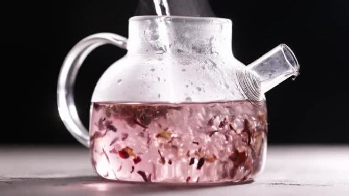 在深色背景下的玻璃透明茶壶中酿造粉红色花朵茶的过程。将热水倒入带茶叶的水壶中。