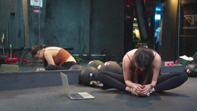 年轻女子通过在健身房的笔记本电脑上观看在线教学课来伸展和锻炼