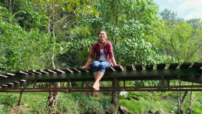 一位美丽的亚洲妇女坐在山林中穿过小溪的木桥上，享受大自然。假日生活方式和冒险。