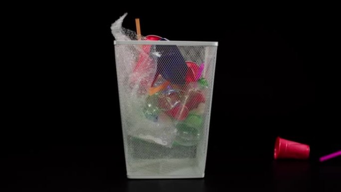 在黑色背景上的空金属垃圾桶中扔塑料垃圾。白色垃圾篮中的玻璃杯，鸡尾酒管，金属罐和瓶子