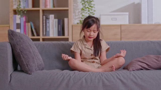 一个亚洲女孩在客厅的灰色沙发上冥想。她穿着休闲装，以使她感到舒适。她对此并不熟悉，但是温暖的地方可以