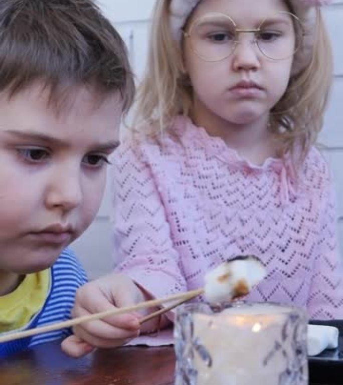 垂直视频。兄妹少年在大蜡烛上用棍子烤棉花糖。那个女孩着火了棉花糖，她被扑灭了