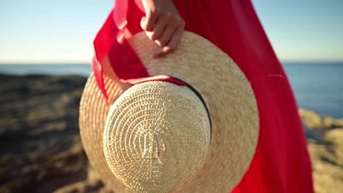 一位穿着红色连衣裙的年轻白人妇女手里的特写草帽站在岩石海岸上。浅景深