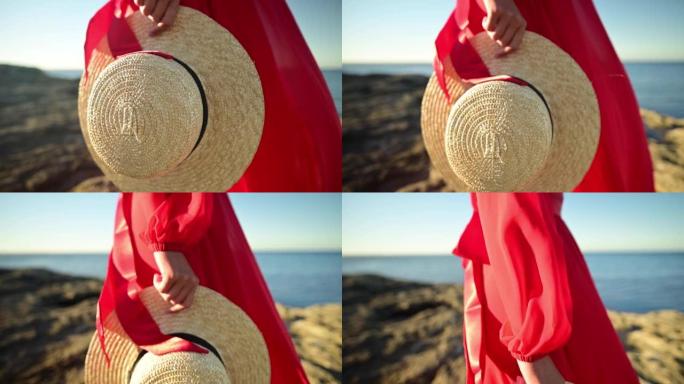 一位穿着红色连衣裙的年轻白人妇女手里的特写草帽站在岩石海岸上。浅景深