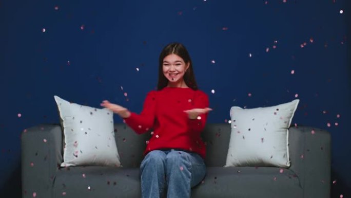 年轻的女孩在闪亮的五彩纸屑下庆祝节日，坐在沙发上的火花或闪光。节日网上商店销售