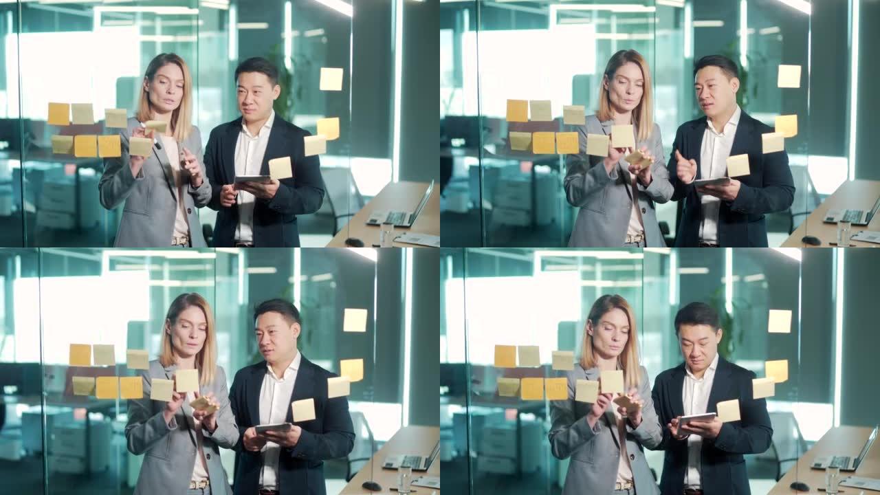 办公室的团队合作会议。两个快乐的亚洲商业同事团队在张贴便签纸上写着便签纸看玻璃