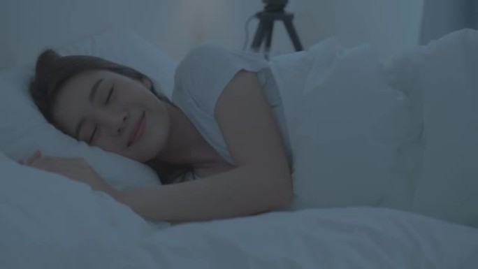 穿着睡衣的亚洲美丽舒适女孩在黑暗的夜晚睡在卧室里。迷人的年轻女子躺在床上，用舒适的枕头和毯子感到幸福