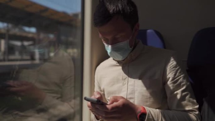 在冠状病毒大流行期间，一名白人男子戴着口罩坐在火车车厢里，使用智能手机。旅游与科技主题