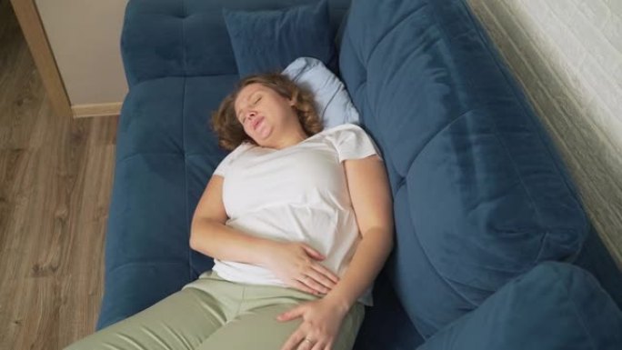 穿着白色t恤的女人躺在蓝色沙发上，用手托着肚子。她用手抚摸肚子，以免受伤。月经，怀孕，腹泻，暴食。脸