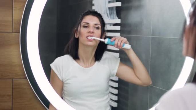 可爱的自信的30岁黑发女人穿着白色t恤在浴室镜子前刷牙