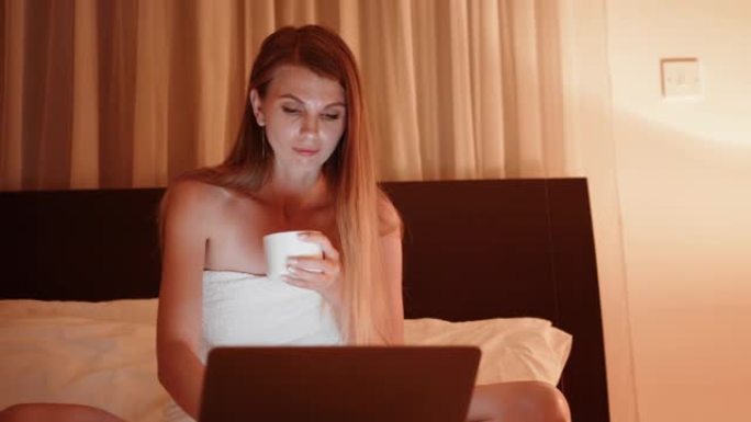疲倦的女人晚上在床上使用笔记本电脑。女士用现代笔记本电脑用浴巾包裹，晚上喝热饮料。坐在酒店房间舒适床