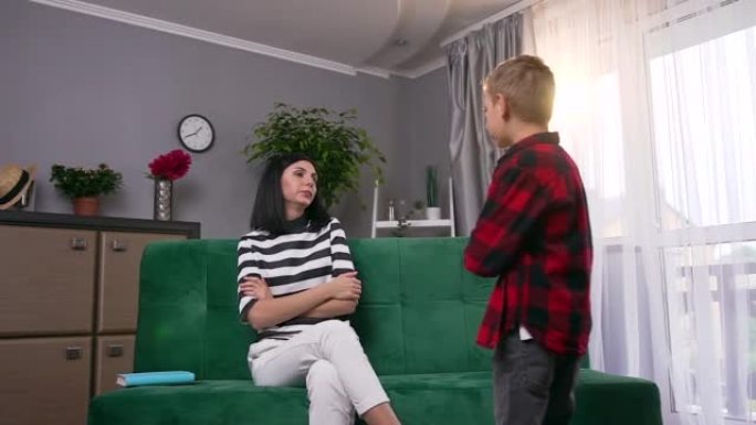 紧张的愤怒的年轻黑发女人坐在家里的客厅沙发上，与愤怒的8岁儿子来回争吵