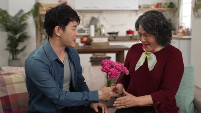 快乐的亚洲成年儿子在母亲节用康乃馨花束给母亲带来惊喜。快乐的高级女人在家里的沙发上微笑着拥抱男人