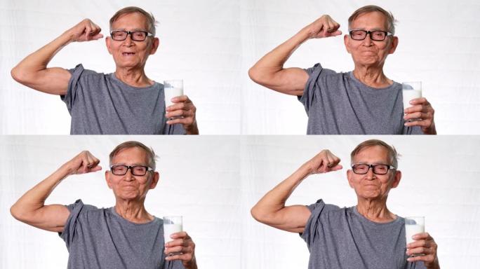 健康的老人拿着一杯牛奶，一边炫耀自己的肌肉，一边在工作室的白色背景上自豪地微笑。