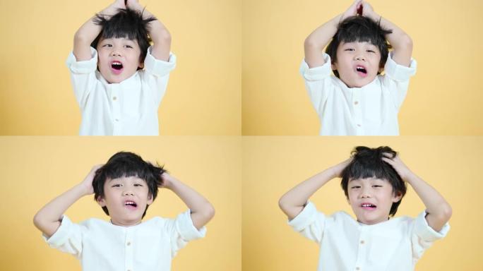 可爱的亚洲男孩的特写肖像表现出兴奋，玩得开心，举起手，来回摩擦头发。站在柔和的黄色墙壁颜色背景上。工