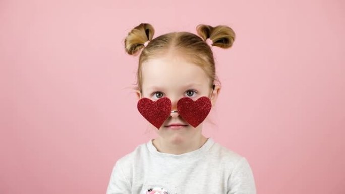有趣的金发小女孩微笑着玩粉色背景的红色心形太阳镜。情人节或母亲节概念