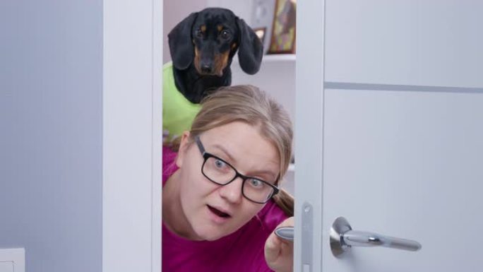 戴着眼镜和黑发腊肠犬的年轻金发女子打开白色的门，偷听好奇的特写镜头